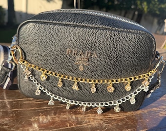 May include: Black leather crossbody bag with a gold chain and silver chain with decorative charms. The bag has a zipper closure and the word "PRADA" in gold lettering. The bag is sitting on a wooden surface.