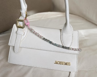 May include: A white leather handbag with a top handle and a flap closure. The bag features gold-colored hardware and the brand name "JACQUEMUS" in gold lettering. A beaded strap with the words "LOVE YOU MORE" is attached.