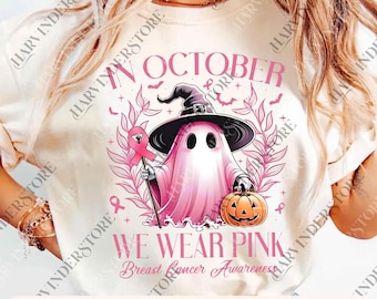 May include: A cream-colored t-shirt featuring a pink ghost wearing a witch hat and holding a pink ribbon and a jack-o'-lantern. The text reads "In October We Wear Pink Breast Cancer Awareness". The design includes bats and pink ribbons.
