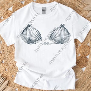 May include: White t-shirt featuring a seashell bra design. The graphic is a detailed illustration of a seashell bikini top, with dark blue lines and bow details. The shirt is a casual, everyday wear item.