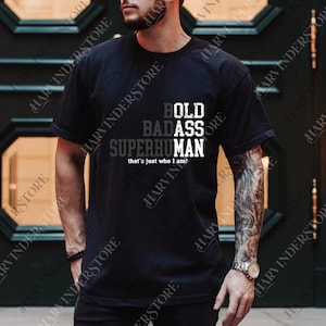 Può includere: T-shirt nero con la scritta "BOLD BADASS SUPERHUMAN" in lettere bianche e grigie. La frase "that's just who I am!" è sotto. La maglietta è a girocollo.