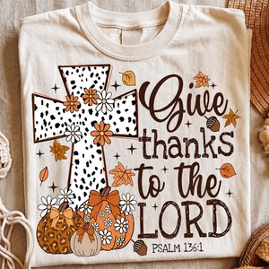May include: A cream-colored t-shirt featuring a graphic of a cross with a black and white dalmatian print, decorated with orange and white flowers. Below are three pumpkins with leopard and floral patterns. Text reads: Give thanks to the LORD PSALM 136:1.
