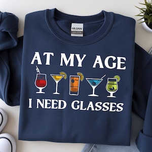 May include: A navy blue crewneck sweatshirt featuring the white text AT MY AGE I NEED GLASSES. Between the two lines of text are five colourful illustrations of various cocktail glasses, including a red drink, a yellow martini, and a green drink.