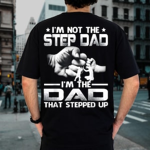 May include: Black t-shirt with white text and a graphic of two hands clasped together. The text reads "I'M NOT THE STEP DAD I'M THE DAD THAT STEPPED UP". The shirt is a crew neck and has short sleeves.