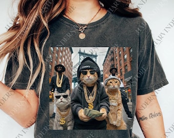 May include: Dark gray t-shirt featuring a graphic of four cats dressed as rappers in a city setting. The cats wear gold chains, sunglasses, and hoodies. One cat holds cash. The word "Canpary" is on a hoodie.