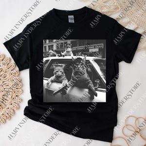 A black t-shirt with a black and white photograph of two cats sitting in a car, one of them holding a gun.