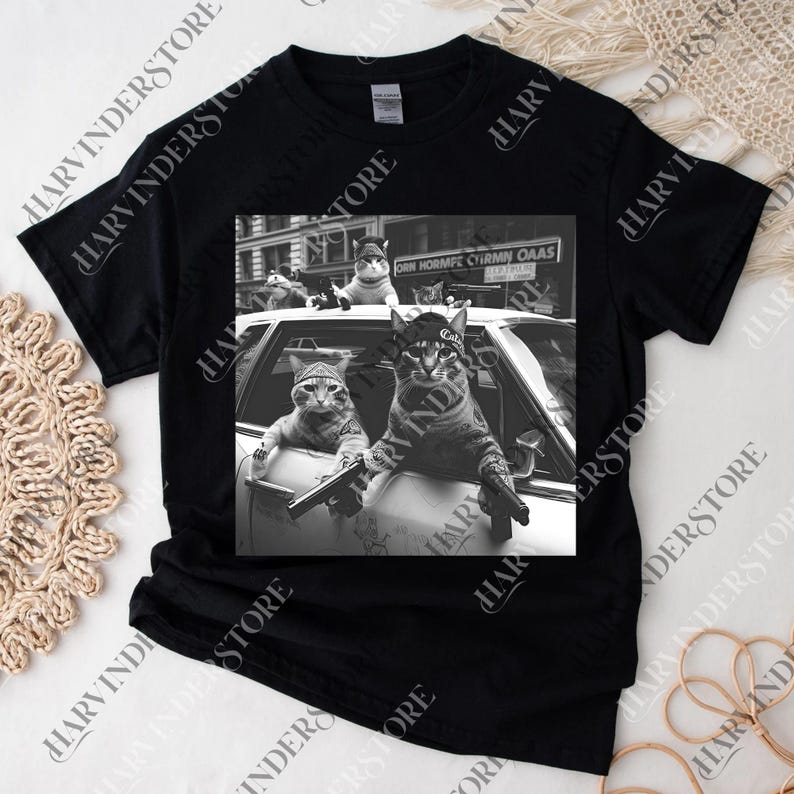 A black t-shirt with a black and white photograph of two cats sitting in a car, one of them holding a gun.