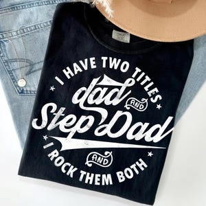 Può includere: Una t-shirt nera con testo bianco dall'aspetto vissuto che recita I HAVE TWO TITLES DAD AND STEP DAD AND I ROCK THEM BOTH. La maglietta presenta un girocollo e un'etichetta del marchio Comfort Colors visibile all'interno del colletto.