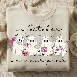 Pode incluir: Uma camisola de gola redonda cor de creme com um gráfico de quatro fantasmas vestidos de enfermeiros com acessórios médicos cor-de-rosa, incluindo uma ligadura, um estetoscópio e uma mala médica. O texto diz: in October we wear pink, com morcegos.