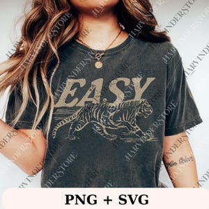 May include: Dark gray t-shirt with the word "EASY" in a distressed font above a tiger illustration. The tiger is in a running pose, with a vintage aesthetic. The shirt is a casual wear item.