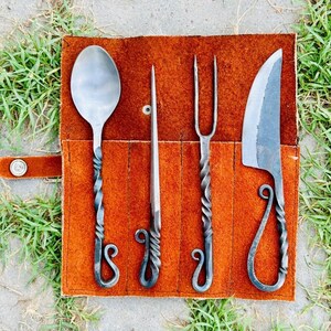May include: A set of four hand-forged cutlery pieces, including a spoon, fork, knife, and a smaller tool, all with twisted handles and decorative loops. The cutlery is displayed in a brown leather roll.