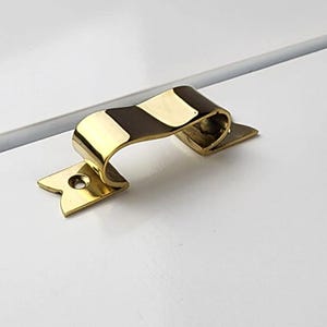 Może przedstawiać: Złoty uchwyt do szuflady z uchwytem w kształcie wstążki i gwiazdkową płytką montażową. Okucia są zamontowane na białej powierzchni, tworząc kontrast między błyszczącym metalem a matowym białym wykończeniem. Projekt jest prosty i elegancki.