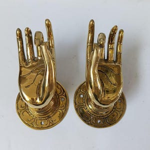 Pode incluir: Um par de puxadores de porta de latão em forma de mãos em posição de oração. Os puxadores são montados em placas redondas com desenhos intrincados.