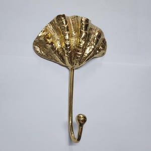 Peut inclure: Un crochet mural doré en forme de coquillage. Le crochet présente un motif texturé et détaillé ressemblant à un coquillage. Le crochet est fixé à une longue tige fine qui se courbe en forme de crochet en bas. Le crochet est en métal.