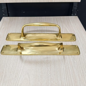 May include: Two brass door pull handles. Each handle has a rectangular backplate with rounded corners and a curved handle. The handles are a shiny gold color and appear to be vintage or antique.