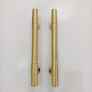 May include: Two vertical door handles made of polished brass with a smooth, cylindrical design. Each handle features a long, straight central bar with slightly thicker, rounded end caps and circular mounting points at the top and bottom.