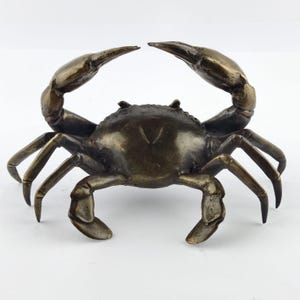 May include: Bronze crab sculpture with raised claws. The crab features a detailed carapace and multiple legs, with a dark bronze finish. The sculpture is set against a white background.