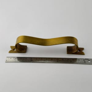 Pode incluir: Um puxador de armário de cor latão com uma alça curva e peças finais decorativas. O puxador tem aproximadamente 35,5 cm de comprimento, medido com uma régua de metal. O hardware está sobre um fundo branco.