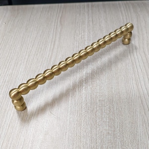 May include: A long, horizontal cabinet handle with a brushed gold finish. The handle features a repeating spherical, bead-like design along its entire length, with two mounting points at each end for attachment to a drawer or cabinet door surface.