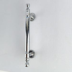 May include: A polished chrome door pull with decorative detailing. The handle is a vertical bar with ornate ends and circular mounting plates. The hardware is designed for a door or cabinet.