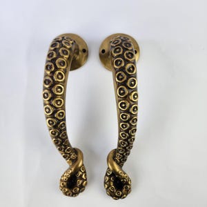 Puede incluir: Dos manijas de puerta de color bronce con un diseño de tentáculo de pulpo. Cada manija presenta una serie de detalles circulares de ventosas a lo largo de su longitud, con una placa de montaje redonda en la parte superior. Las manijas son curvas y tienen una apariencia texturizada.