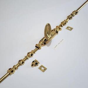 Op de afbeelding: Een goudkleurige, sierlijke deursluiting met een ovale handgreep en decoratieve accenten. Inclusief twee bijpassende montageplaten. De tekst op het label luidt "LARGE OVAL STRIKE".