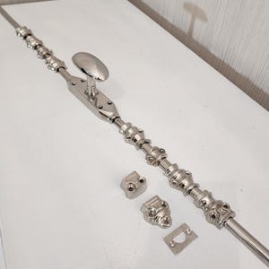 Op de afbeelding: Een gepolijste zilverkleurige deursluiting met een ovale handgreep en decoratieve details. Inclusief montagemateriaal: twee hoekstukken en een sluitplaat. De grendel is ontworpen voor een veilige deursluiting.