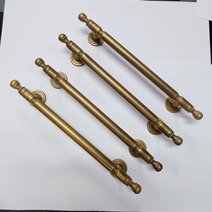 Op de afbeelding: Vier antieke messing deurgrepen. Elke greep heeft een lange, rechte handgreep met decoratieve eindstukken en ronde montageplaten. Het messing heeft een warme, verouderde afwerking, wat een vintage stijl suggereert.
