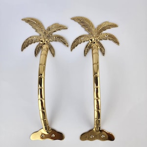 May include: Two gold-colored palm tree door handles. Each handle features a detailed palm frond design at the top and a textured trunk. The handles are curved and have a base with screw holes for mounting. The overall design is tropical.