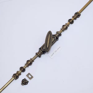 Puede incluir: Un cerrojo de ventana de latón antiguo con un diseño central ovalado y detalles decorativos. Incluye una placa de cierre y un pestillo a juego. El texto "LARGE OVAL STRIPE" es visible en una pequeña etiqueta blanca.