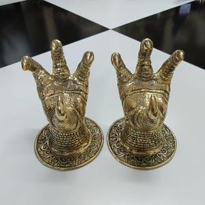 May include: Two brass-coloured decorative rhino-horn sculptures. Each sculpture features a rhino horn and three fingers, mounted on a round base with an ornate, swirling design. The sculptures are displayed on a black and white checkered surface.