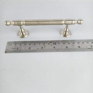 Peut inclure: Une poignée de meuble en métal argenté avec une barre cylindrique et des extrémités décoratives. La poignée mesure environ 15 cm de long. Elle possède deux bases de montage circulaires. Une règle est placée en dessous pour la mesure.