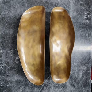 Könnte beinhalten: Zwei goldfarbene, gebogene Metallschalen oder -schalen. Die Oberflächen haben eine polierte, reflektierende Qualität mit subtilen Farbvariationen. Die Formen sind organisch und asymmetrisch, was auf ein dekoratives oder funktionales Design hindeutet.