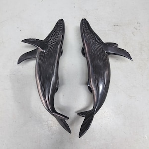 May include: A pair of dark bronze whale-shaped door handles. Each handle has a detailed whale design, with fins, tail, and textured body. The handles are mounted on a grey surface, highlighting their sculptural form and decorative appeal.
