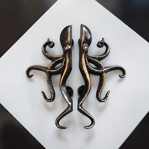 Peut inclure: Deux poignées de porte en forme de poulpe de couleur bronze. Chaque poignée présente un corps central avec des tentacules incurvées. Les poignées sont montées sur une surface à carreaux blancs et noirs.