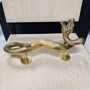 Puede incluir: Un tirador de puerta de latón pulido con forma de serpiente de cascabel enroscada. La serpiente presenta escamas detalladas, la cabeza levantada con la boca abierta mostrando los colmillos y un cascabel en la cola. Montado sobre dos bases circulares.