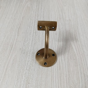 Puede incluir: Un soporte de pasamanos de latón con un soporte curvo y una base circular. El soporte tiene una parte superior plana con dos orificios de montaje y una base con cuatro orificios. El metal tiene un aspecto cálido y envejecido.