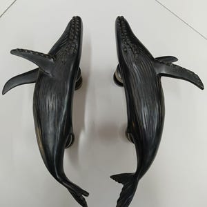 May include: Two dark bronze whale-shaped door handles. Each handle features a detailed carving of a whale, including fins, tail, and textured skin. The handles are mounted on circular backplates.