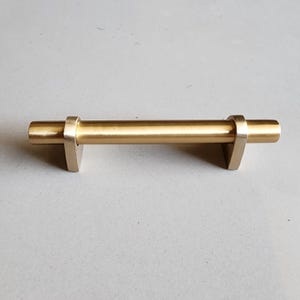 May include: A gold-toned metal cabinet pull with a rectangular shape and rounded edges. The pull has a smooth, polished finish.