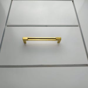 May include: A gold-colored cabinet pull with a rectangular bar and square end pieces. The pull is mounted on a white cabinet drawer. The hardware has a modern, minimalist design.