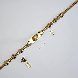 Puede incluir: Un cerrojo de puerta de latón con un centro ovalado y detalles decorativos. El cerrojo va acompañado de dos placas de cierre y una etiqueta que dice "SMALL OVAL". El herraje se muestra sobre una superficie blanca.