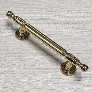 May include: A gold-colored cabinet pull with a cylindrical bar and decorative finials. The pull has circular mounting plates on each end. The hardware is made of metal and has a polished finish.