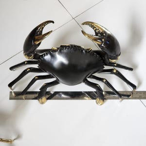 May include: A decorative crab sculpture in black with gold accents on the claws and joints. The crab is positioned on a white surface, showcasing its detailed design. The sculpture is approximately 38 cm wide.