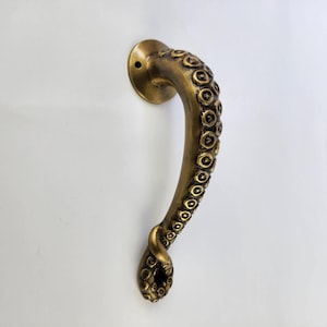 May include: A bronze-colored door handle shaped like an octopus tentacle. The handle features a circular mounting plate and is detailed with suction cups along its curved length. The handle is mounted on a white surface.