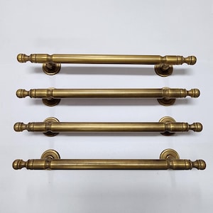 May include: Four antique brass door pulls. Each pull features a cylindrical bar with decorative end pieces and circular mounting plates. The brass has a warm, aged patina. The pulls are arranged horizontally against a white background.
