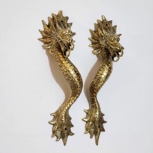 May include: Two antique brass dragon door handles. Each handle features a detailed dragon head, a scaled body, and clawed feet. The handles are a warm gold color and are set against a white background. These decorative handles would be a unique addition to any door.