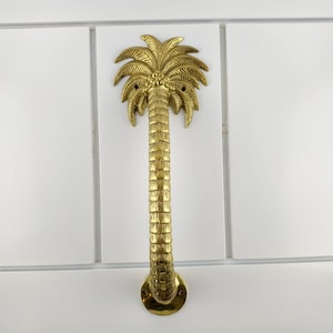 Może przedstawiać: Złoty uchwyt do drzwi w kształcie palmy. Uchwyt jest wykonany z metalu i ma szczegółowy wzór, w tym liście palmowe i pień. Uchwyt jest zamontowany na białej powierzchni.