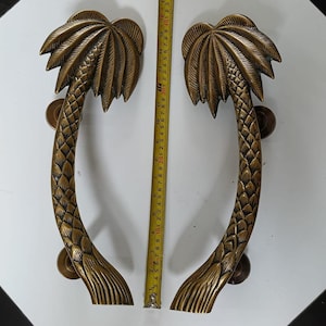 Puede incluir: Dos manijas de puerta de color bronce, cada una con forma de palmera. Las manijas tienen un diseño curvo con una fronda de palmera en la parte superior y un tronco texturizado. Cada manija mide aproximadamente 30 cm de largo e incluye herrajes de montaje.
