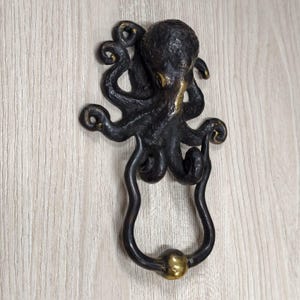 Op de afbeelding: Bronzen octopusdeurknocker met een gedetailleerd, gestructureerd ontwerp. De octopus heeft acht gekrulde tentakels en een ronde kop. De handgreep van de knocker heeft een gebogen vorm met een messingkleurige bal aan de onderkant. Gemonteerd op een licht houten oppervlak.