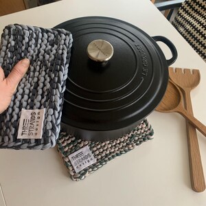 May include: A black Le Creuset Dutch oven with a silver knob, alongside two handmade pot holders in grey and green hues. Wooden cooking utensils are also visible. The pot holders feature a label reading "THREE STRANDS design."